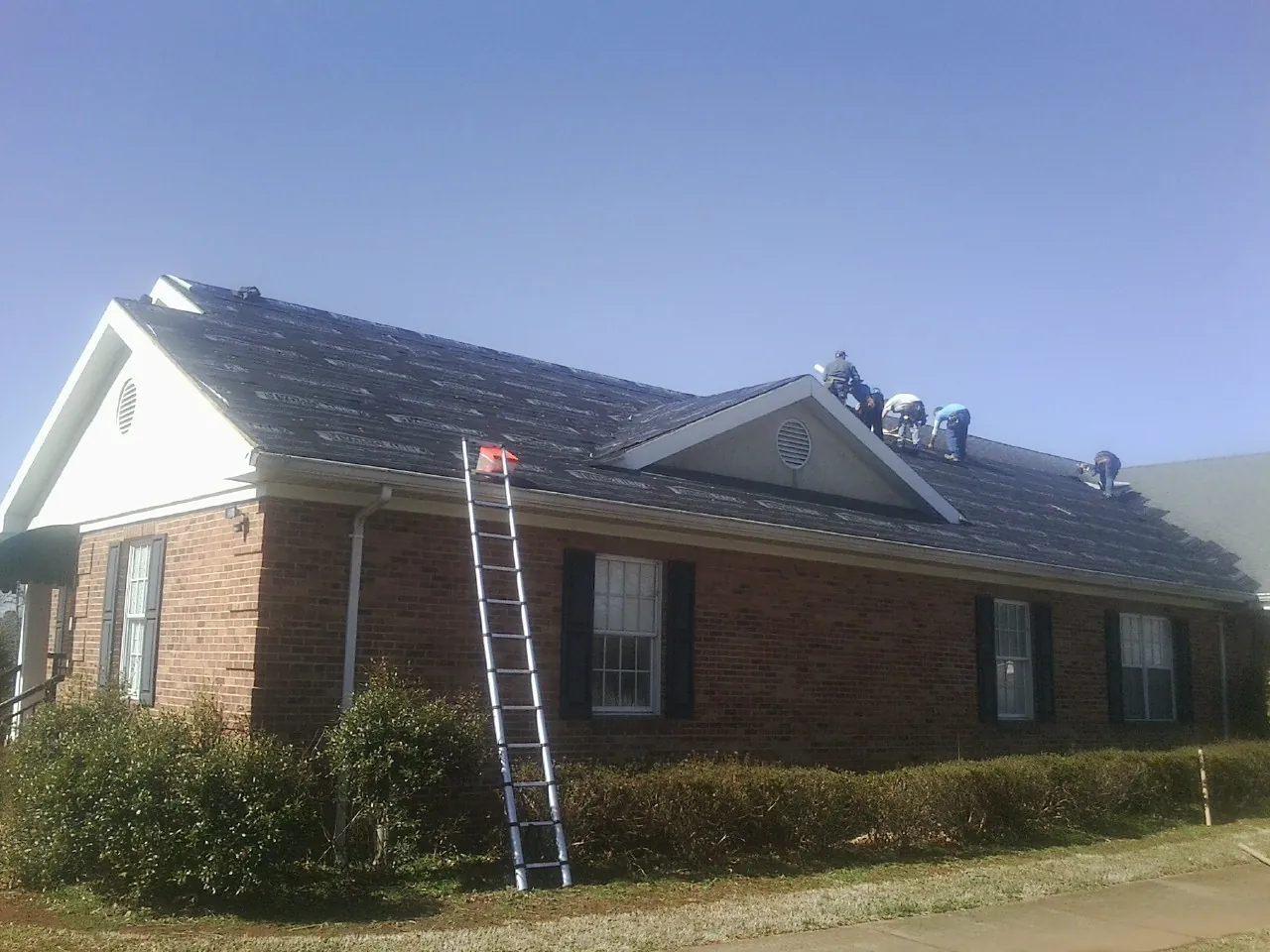 Crew tearing shingles off a brick ranch roof with a ladder set — Emergency Roof Repair work by our crew in Sigourney
