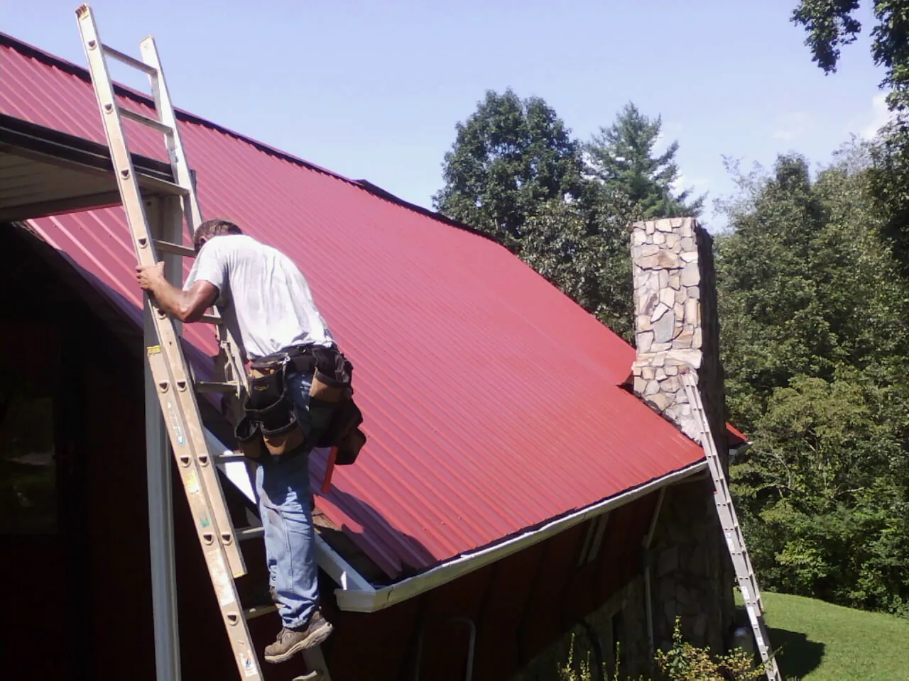 Roofer on a ladder beside a red metal roof and stone chimney — Soffit & Fascia Repair by Roofing Brothers in Sigourney